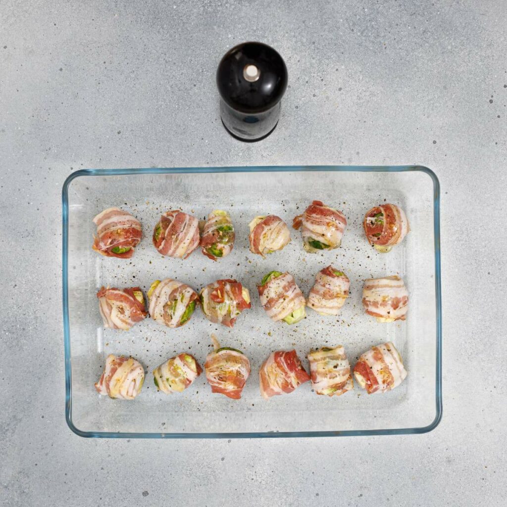 A glass baking dish with rows of bacon-wrapped vegetables, sprinkled with pepper, sits on a gray countertop with a black pepper grinder above it.