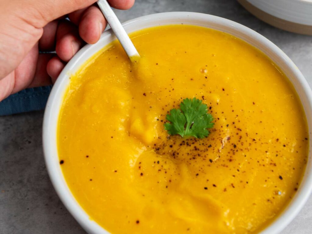 A hand holding a spoon in a bowl of creamy squash soup, garnished with black pepper and a sprig of cilantro.