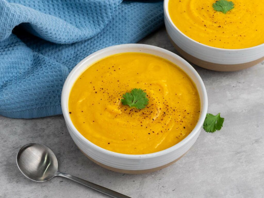 Two bowls of squash soup, each garnished with a cilantro leaf and black pepper, sit on a gray surface.