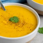A bowl of creamy squash soup garnished with black pepper and a cilantro leaf, with a spoon inside.
