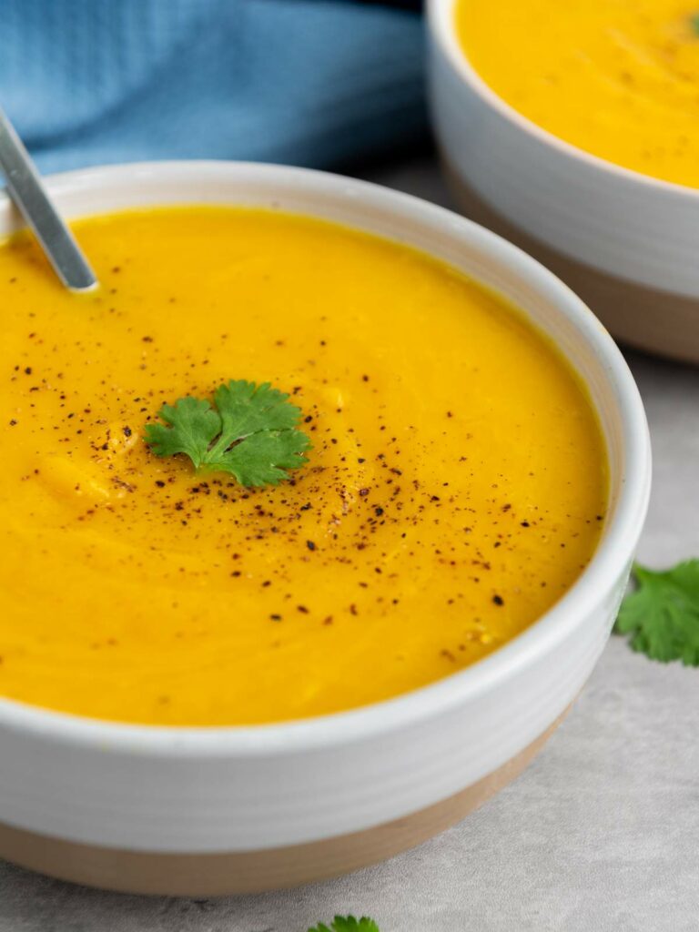A bowl of creamy squash soup garnished with black pepper and a cilantro leaf, with a spoon inside.