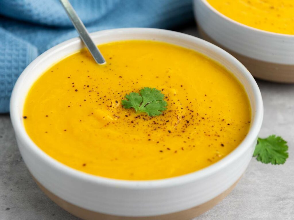 A bowl of creamy squash soup garnished with a cilantro leaf and black pepper, with a spoon in the bowl.