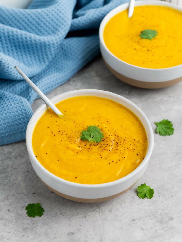 Two bowls of creamy orange soup garnished with cilantro leaves and black pepper, each with a spoon.
