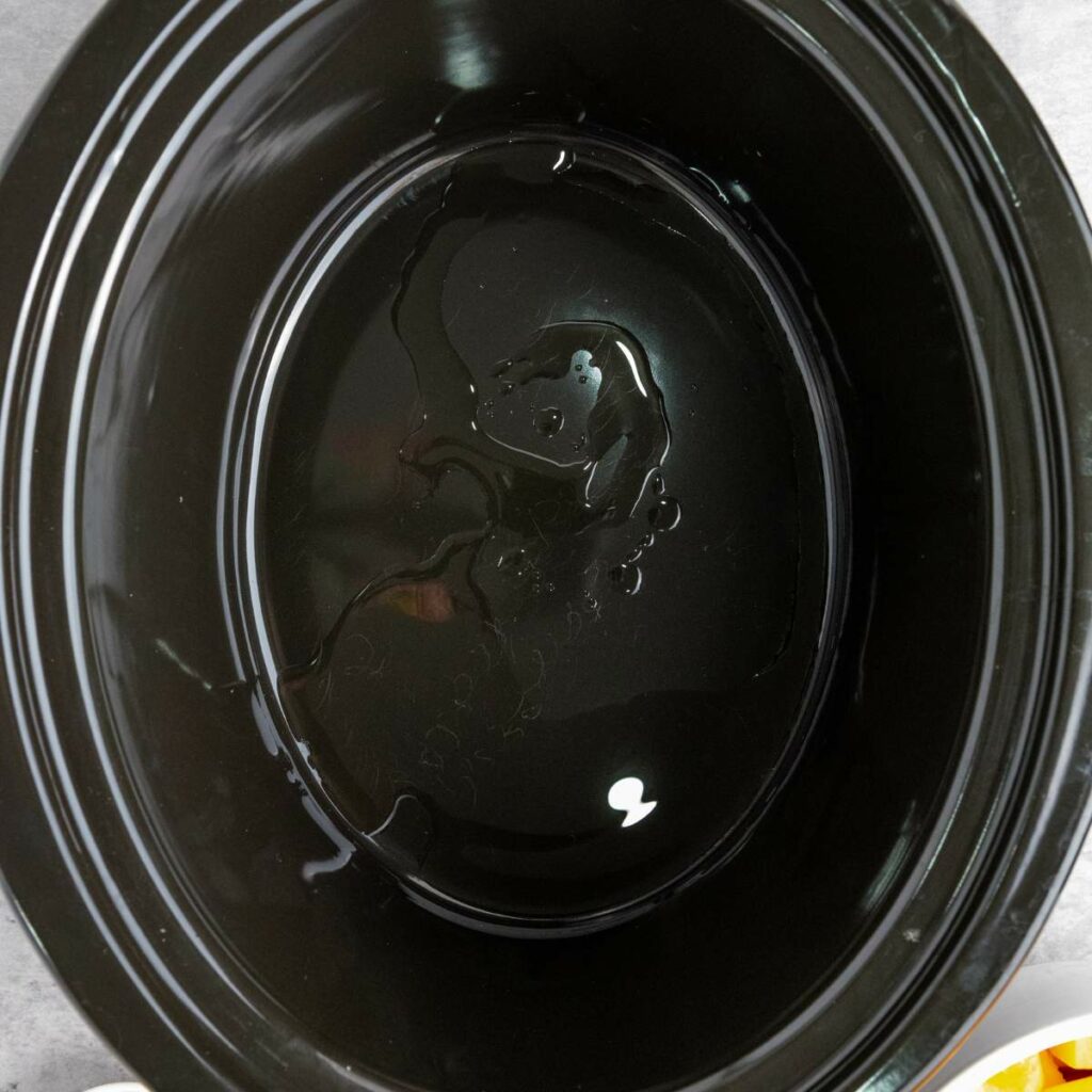A black slow cooker with a small amount of oil at the bottom on a gray surface.