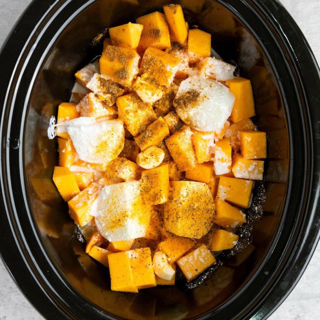 Cubed butternut squash, coconut milk, and spices in a black slow cooker, ready to be cooked.
