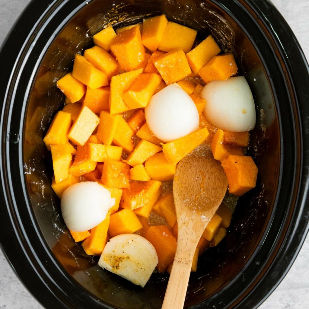 Chopped butternut squash and quartered onions in a slow cooker with a wooden spoon, ready for cooking.