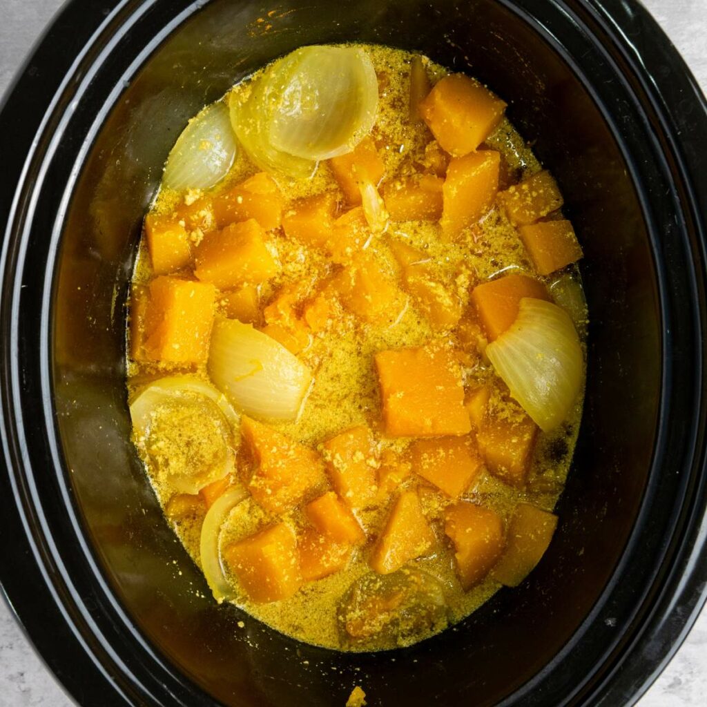 Chopped butternut squash and onion pieces in a slow cooker with broth and seasonings.