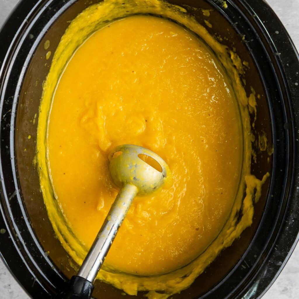 A slow cooker filled with smooth yellow soup and an immersion blender resting inside.