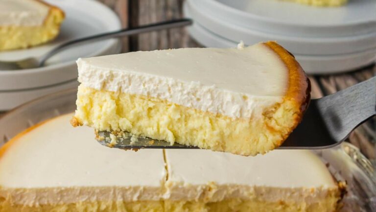 A slice of cheesecake with a creamy topping being lifted from a whole cheesecake, with plates and a fork in the background.