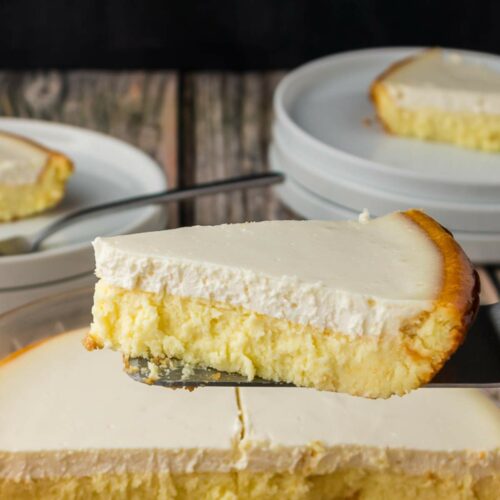 A slice of cheesecake with a creamy top layer is being lifted from a pan, with more slices and stacked white plates in the background.