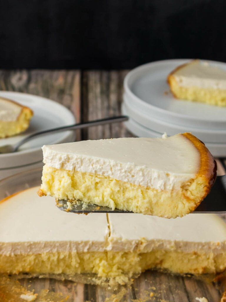 A slice of cheesecake with a creamy top layer is being lifted from a pan, with more slices and stacked white plates in the background.