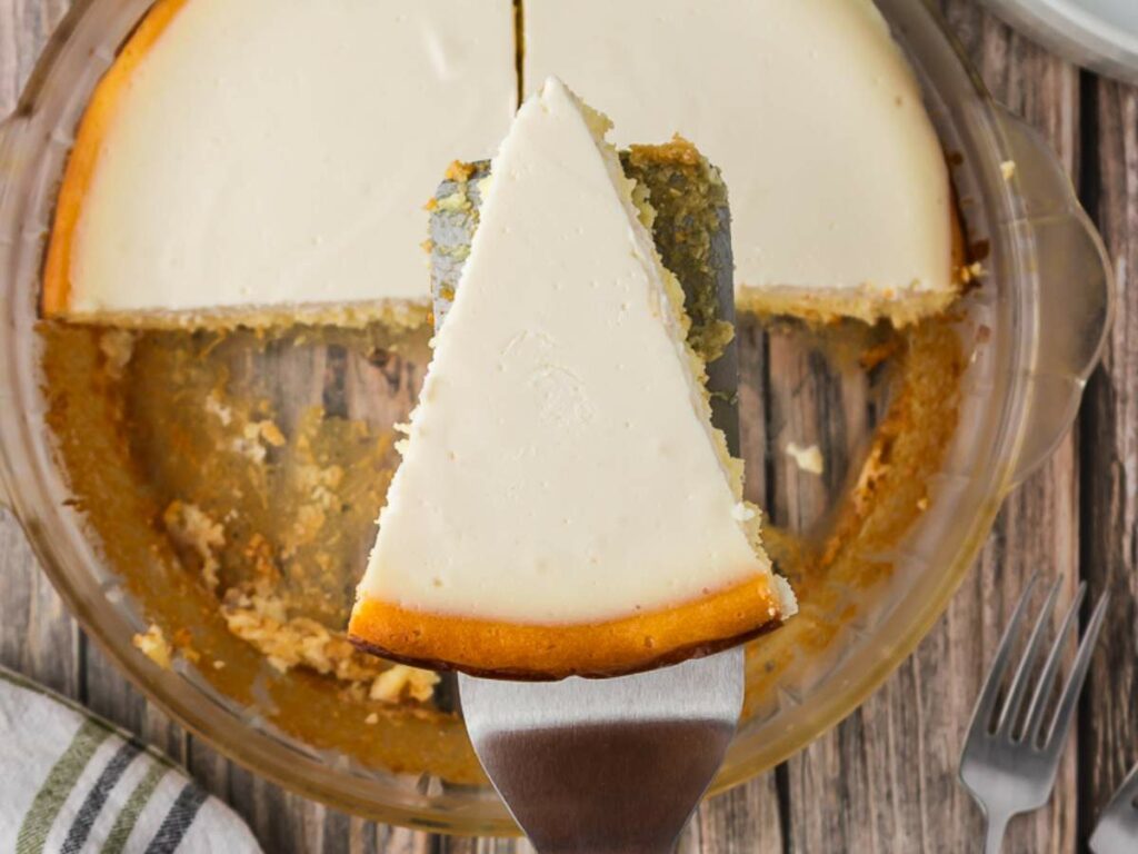 A slice of cheesecake being lifted from a glass pie dish, with the remaining cheesecake and crumbs visible on a wooden surface.