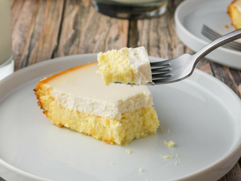 A slice of cheesecake with a creamy topping on a white plate; a fork holds a bite of cake above the slice.