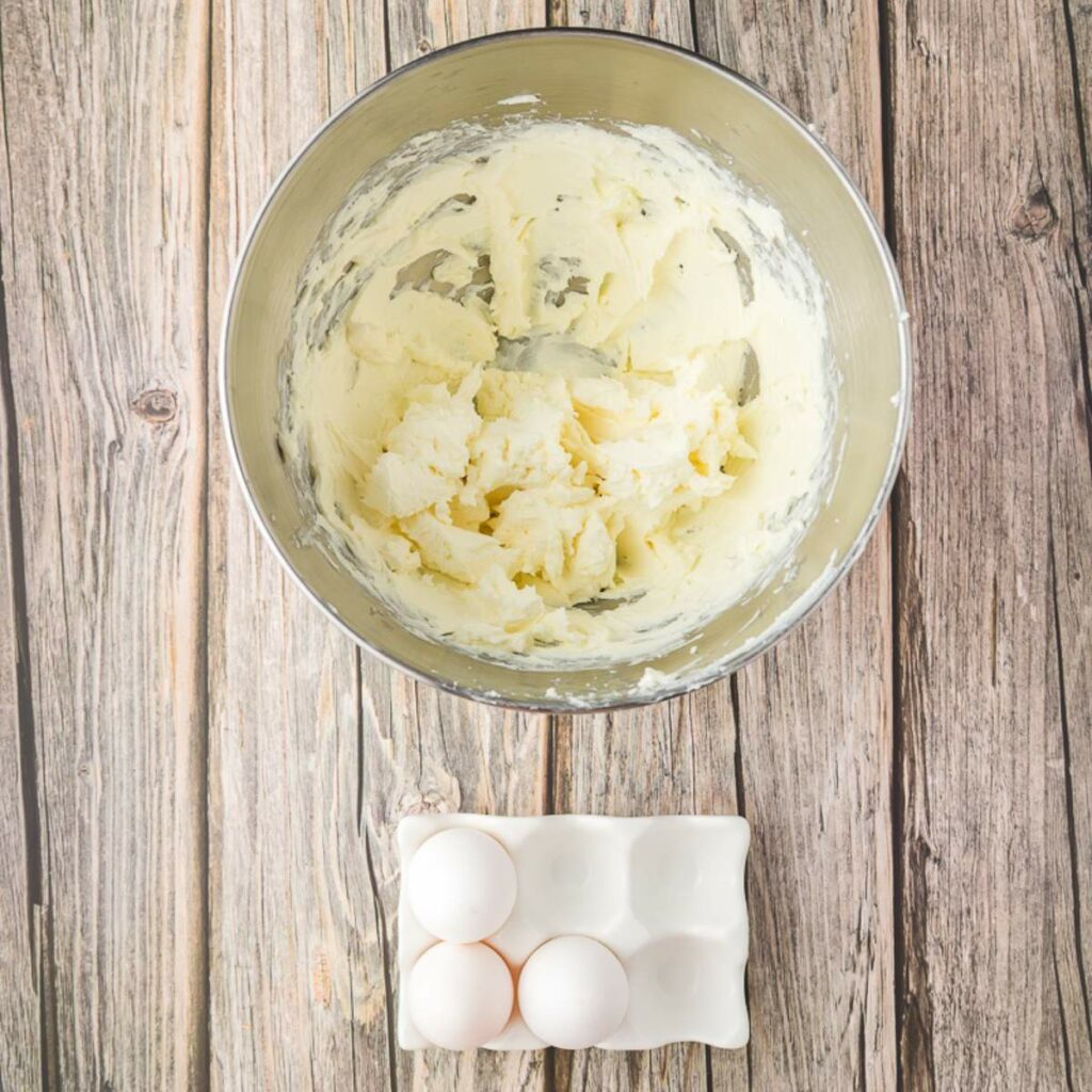 A metal mixing bowl with creamed butter sits on a wooden surface above a ceramic tray holding three eggs.