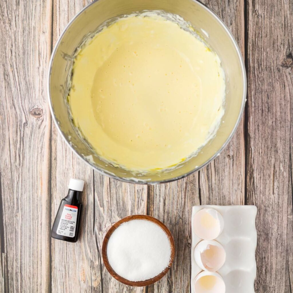 A mixing bowl with batter, a small bottle of vanilla extract, a bowl of sugar, and cracked eggs in a tray on a wooden surface.