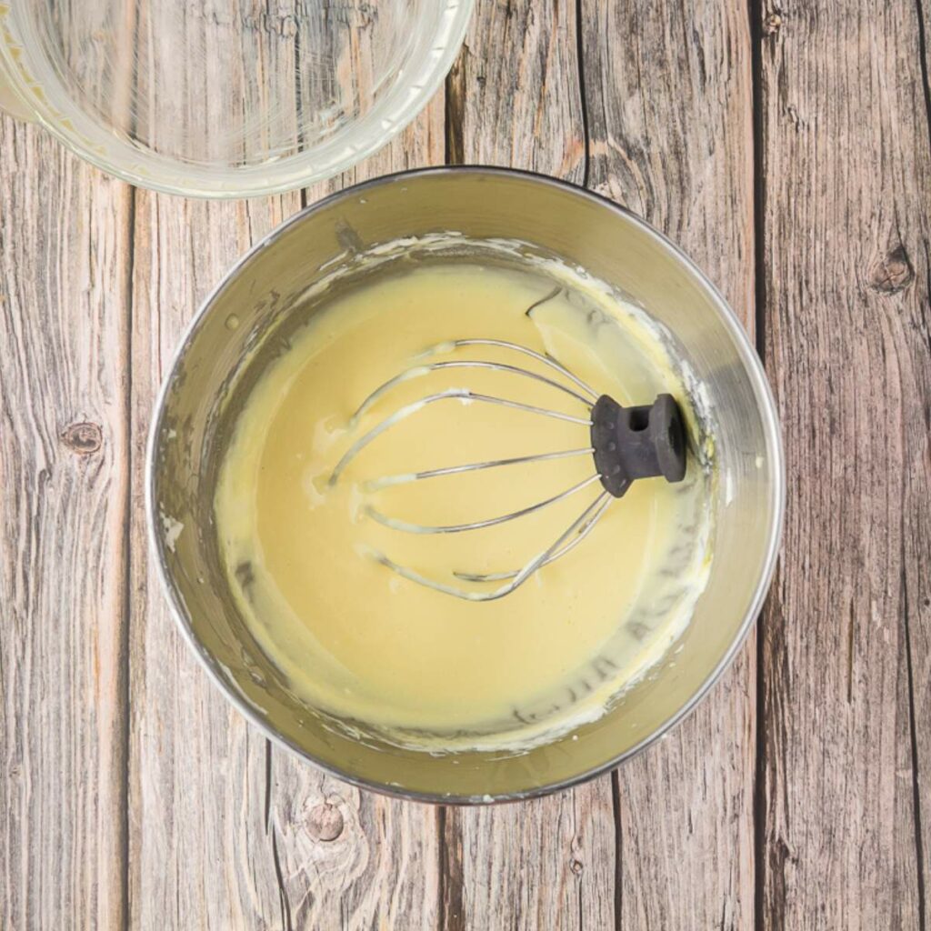 A metal mixing bowl with a whisk attachment sits on a wooden surface, containing a pale yellow mixture.