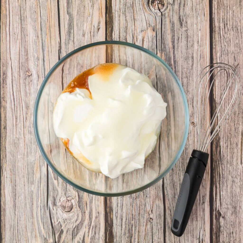 A glass bowl with whipped cream and a brown liquid, possibly a syrup, on a wooden surface next to a metal whisk with a black handle.