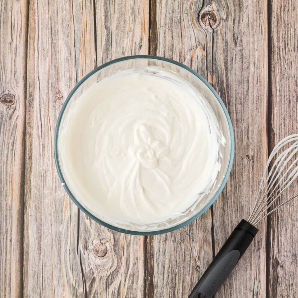 A glass bowl filled with whipped cream sits on a wooden surface next to a metal whisk with a black handle.