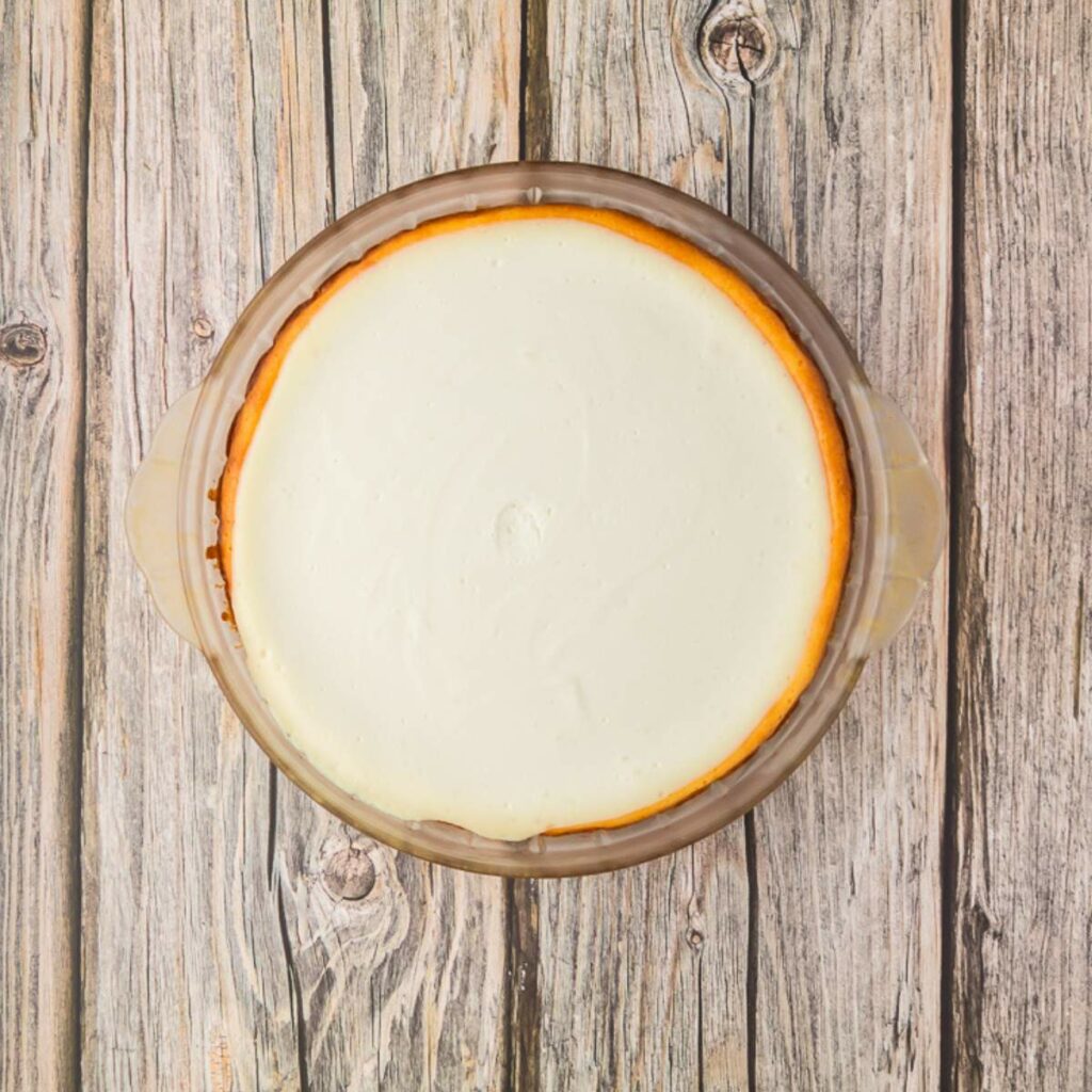 A plain cheesecake in a round glass baking dish placed on a wooden surface, viewed from above.