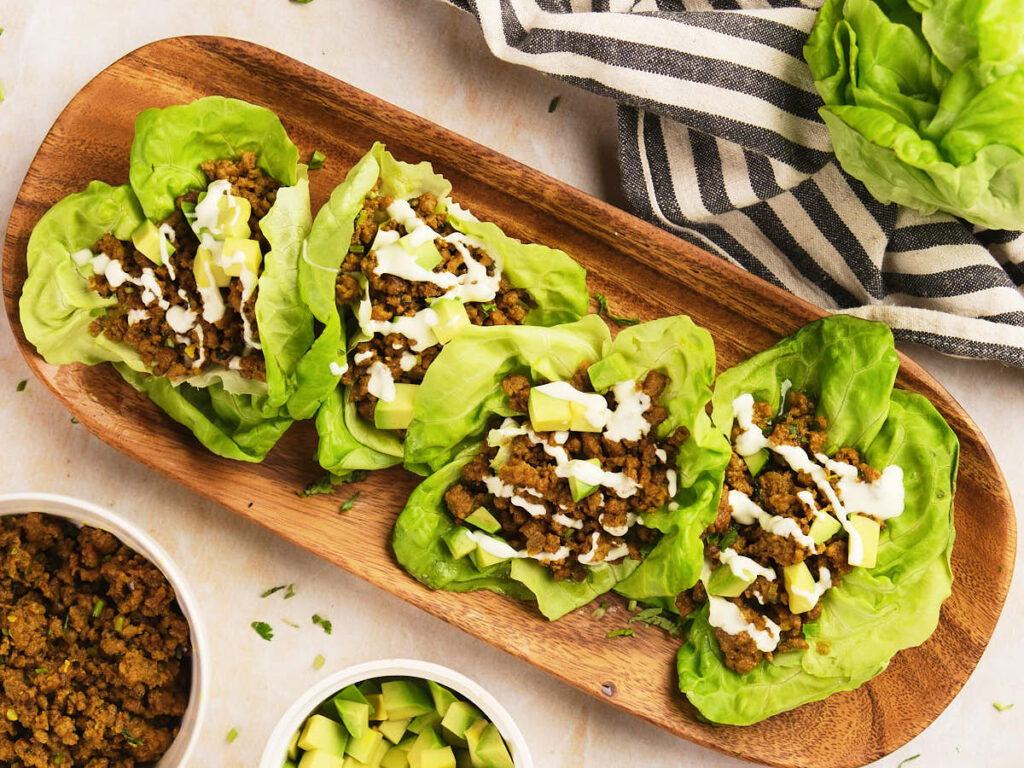 Four lettuce wraps filled with seasoned ground meat, diced avocado, and a drizzle of white sauce.