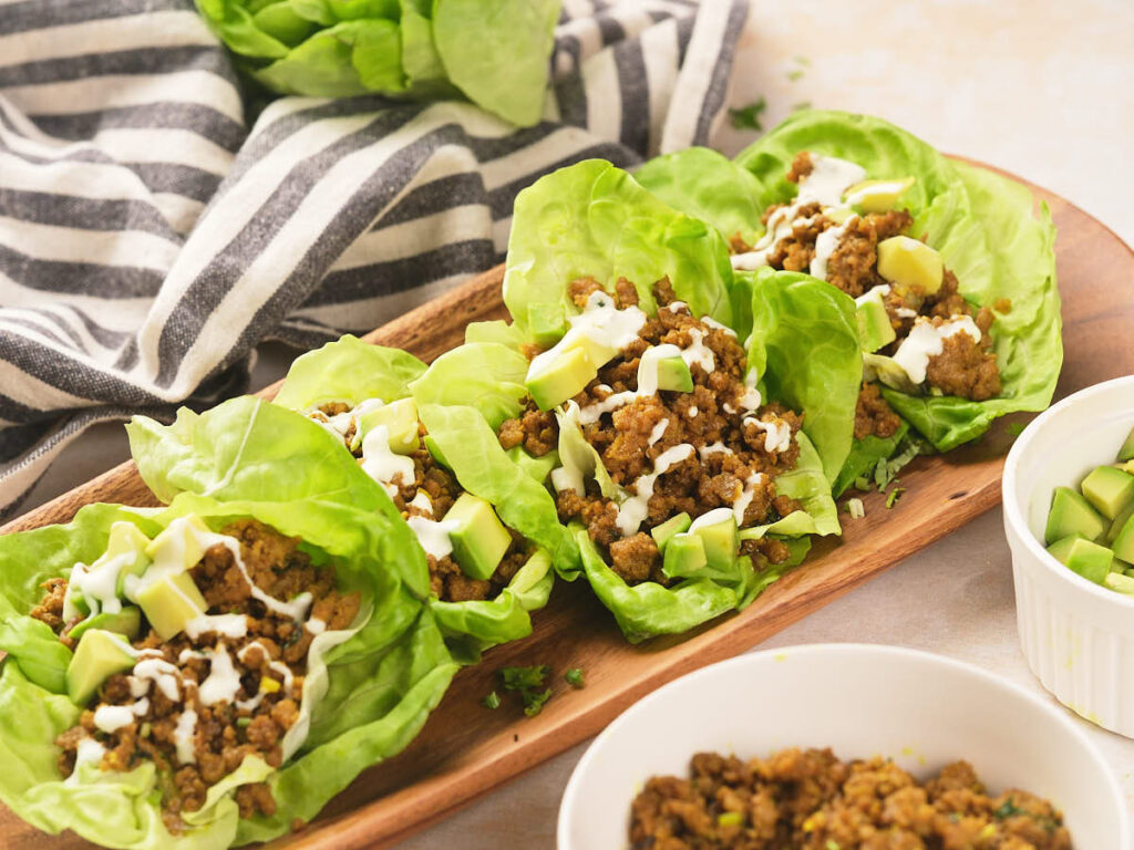 Four lettuce leaves filled with seasoned ground beef, diced avocado, and drizzled with a white sauce are arranged on a wooden tray.