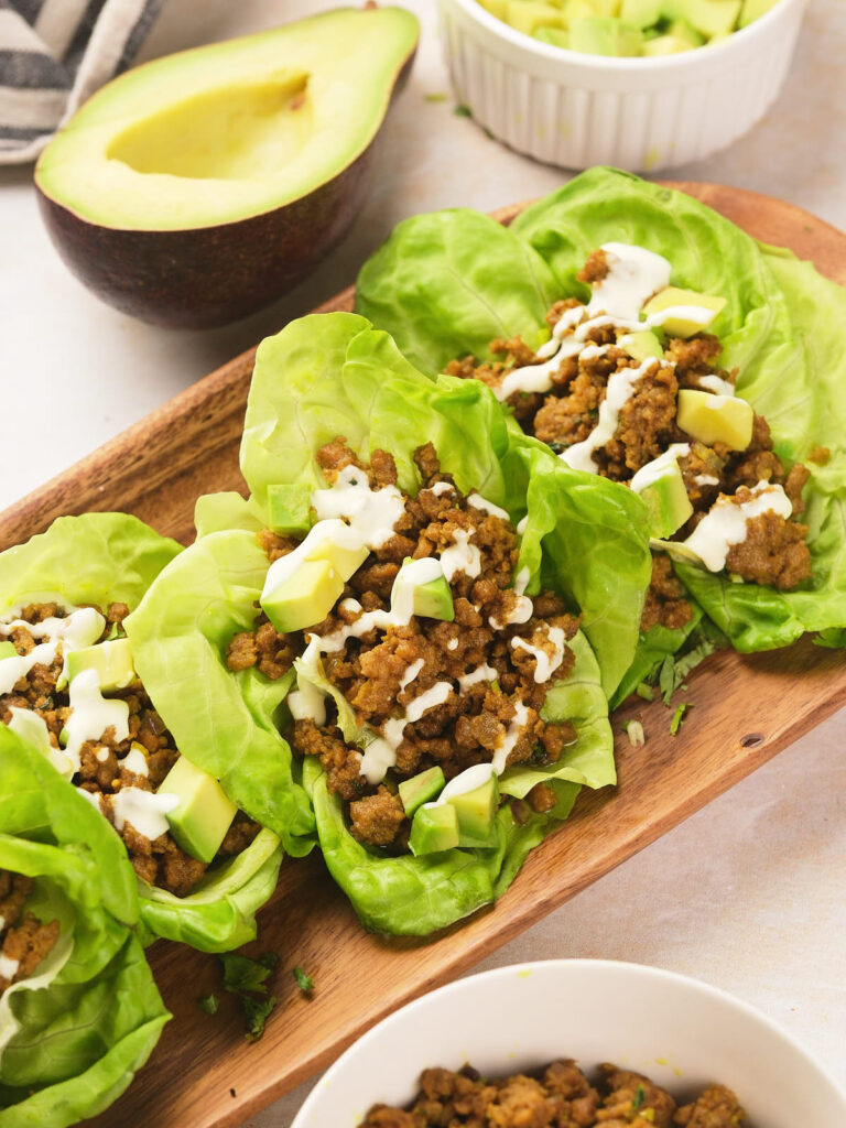 Three lettuce leaves filled with seasoned ground meat, diced avocado, and a drizzle of white sauce.