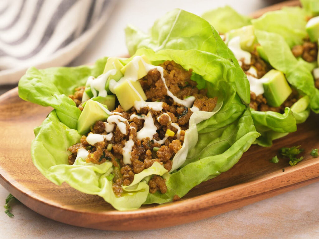 Lettuce wraps filled with seasoned ground beef, diced avocado, and drizzled with a white sauce, served on a wooden plate.