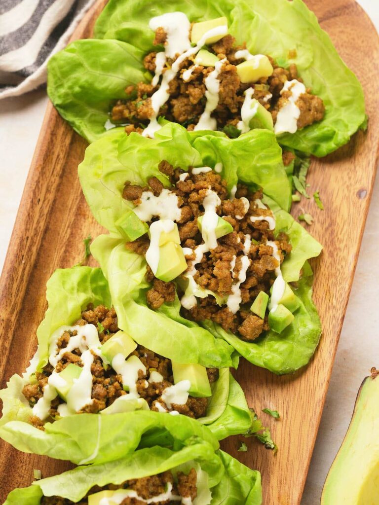 Four lettuce leaves on a wooden tray, each filled with crumbled cooked ground beef, diced avocado, and drizzled with a creamy white sauce.