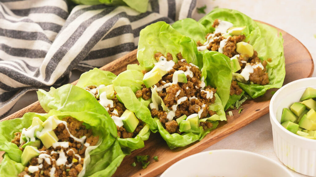 Lettuce leaves filled with seasoned ground meat, diced avocado, and drizzled with white sauce, served on a wooden tray.