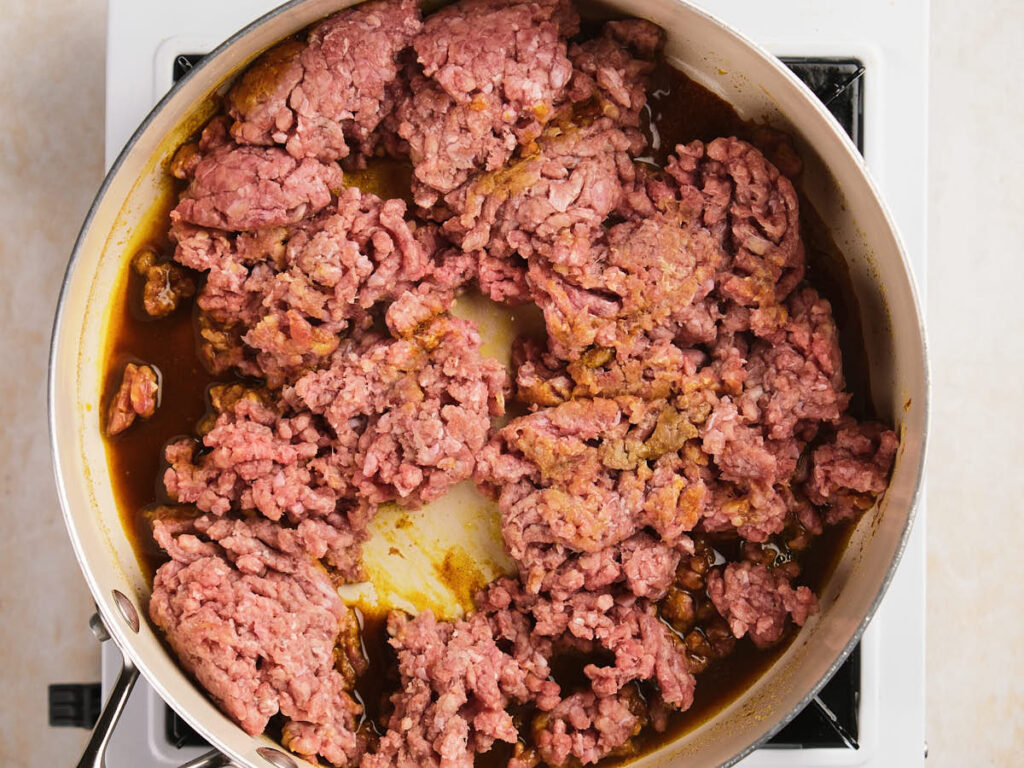Ground beef being cooked in a pan on a stovetop, with some parts browned and some still pink.