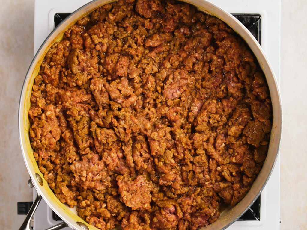A skillet filled with cooked ground meat seasoned with spices sits on a white stovetop.
