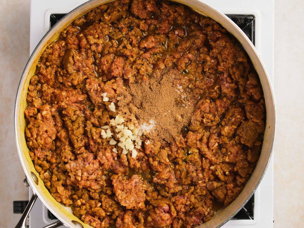 Ground meat cooking in a skillet on a stove, with chopped garlic and spices added on top, before being stirred in.