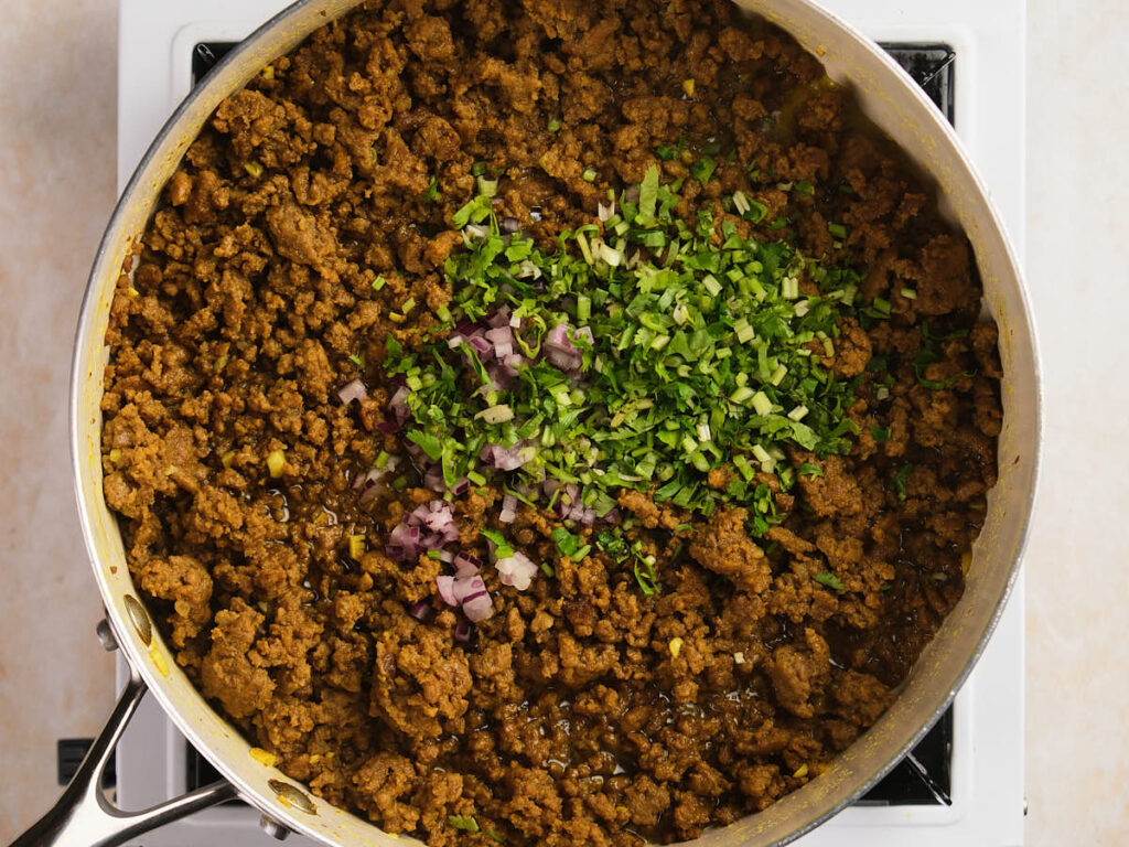 Ground meat cooking in a skillet on a stovetop, topped with chopped herbs and diced onions.