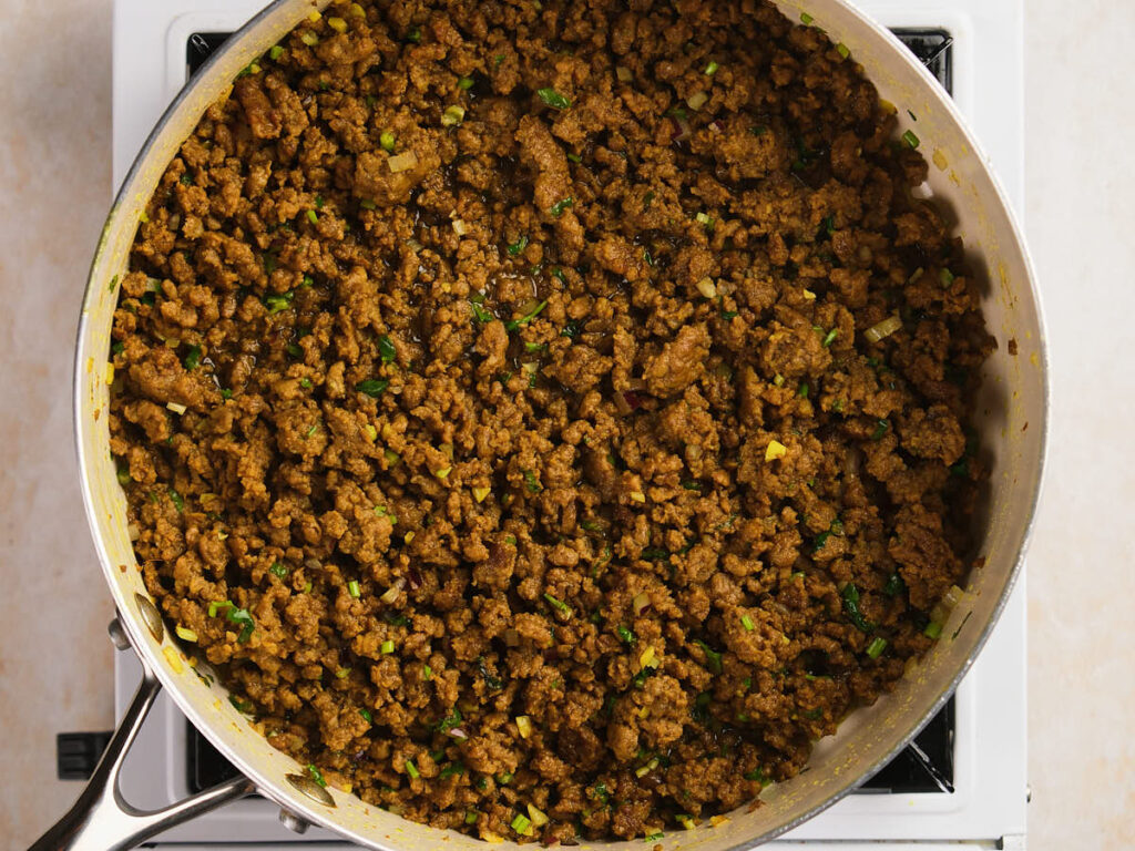 A pan filled with cooked ground beef mixed with chopped herbs and spices sits on a stovetop burner.