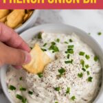 A hand dipping a cracker into a bowl of French onion dip garnished with chopped chives and black pepper.