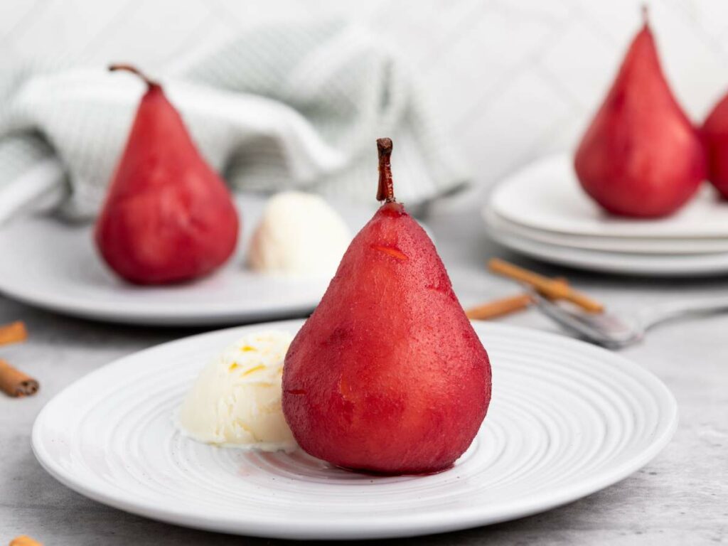 A poached pear with a scoop of vanilla ice cream is served on a white plate, with more pears and cinnamon sticks in the background.