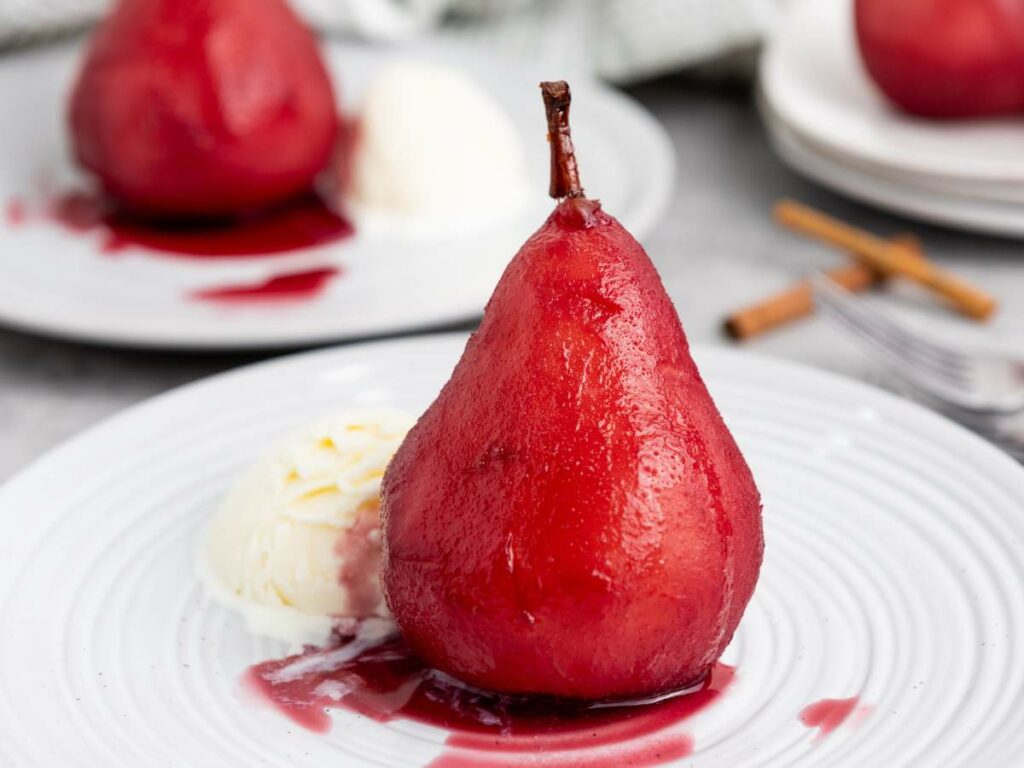 A poached pear in red sauce served with a scoop of vanilla ice cream on a white plate.