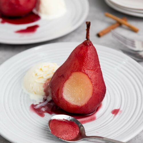 A poached pear served with a scoop of vanilla ice cream on a white plate, with a spoonful of pear and red sauce in the foreground.