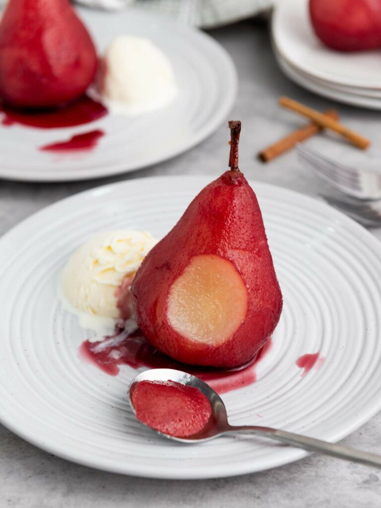 A poached pear served with a scoop of vanilla ice cream on a white plate, with a spoonful of pear and red sauce in the foreground.