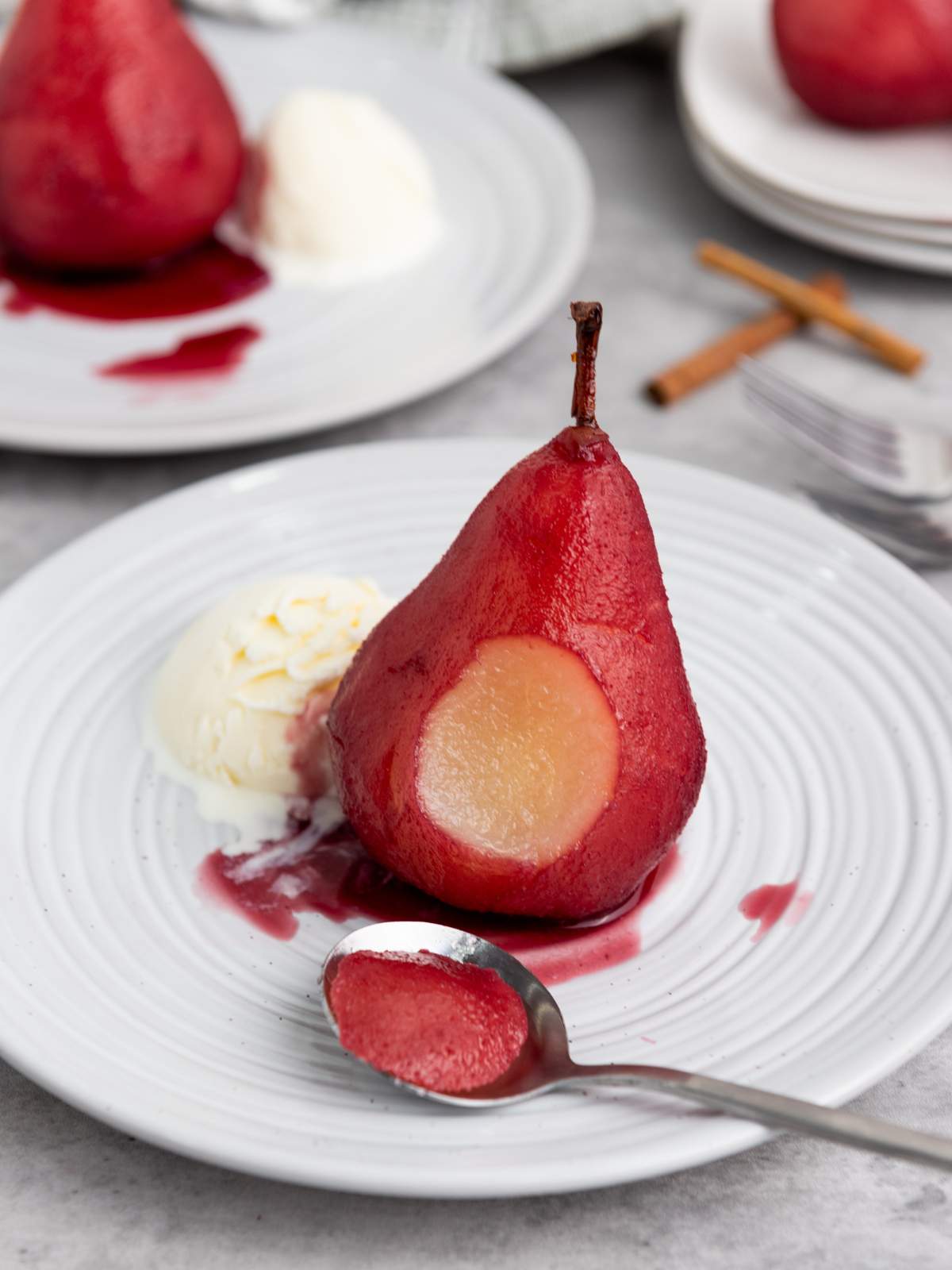A poached pear served with a scoop of vanilla ice cream on a white plate, with a spoonful of pear and red sauce in the foreground.