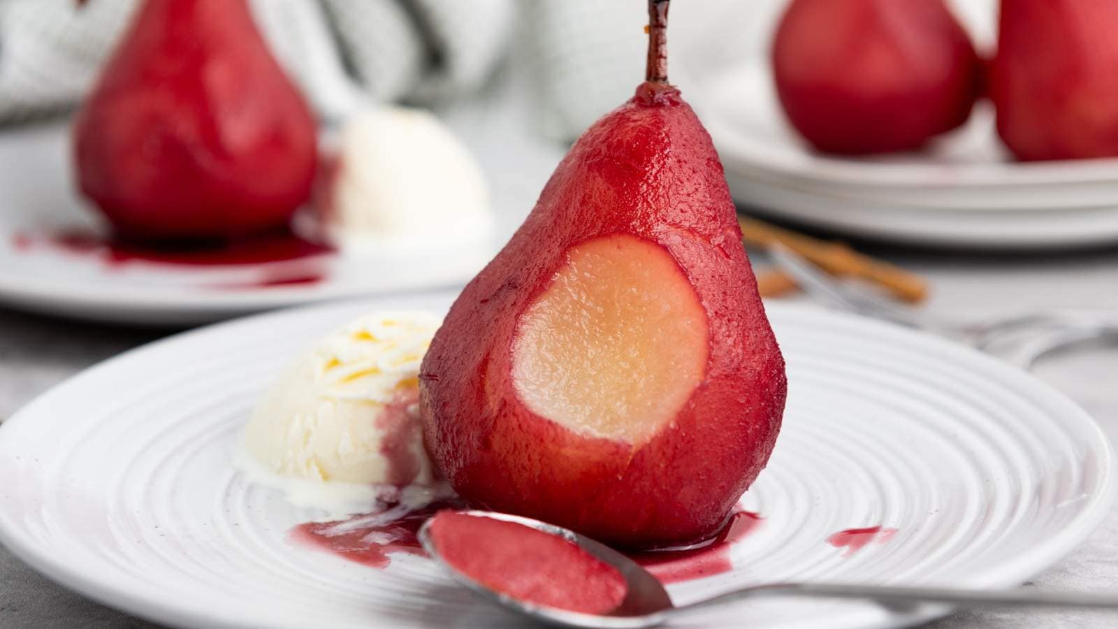 A poached pear with a section scooped, served with a scoop of vanilla ice cream and sauce on a white plate, with a spoon in front.