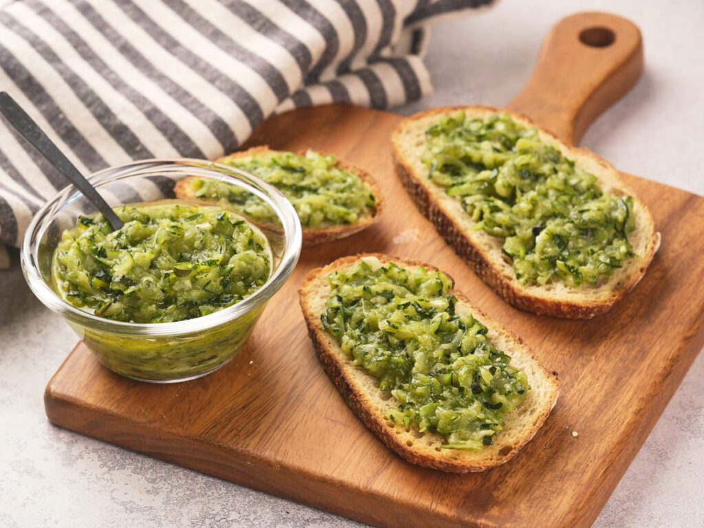 Three slices of bread topped with green zucchini butter are on a wooden board.
