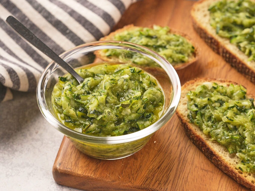 A glass bowl of green butter with a spoon, next to slices of toasted bread.