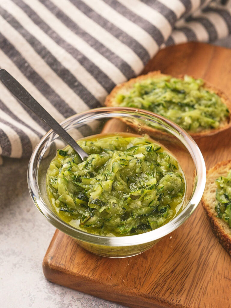 A small glass bowl of grated zucchini with butter and herbs, placed on a wooden board.