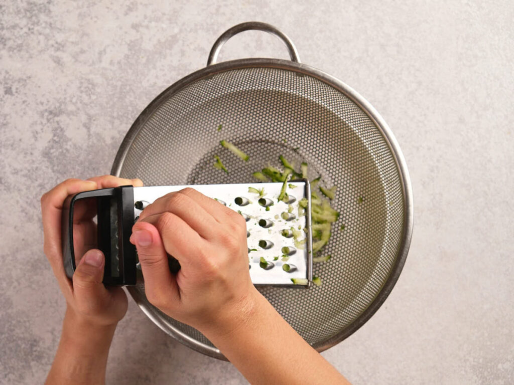 A person grates a zucchini over a metal strainer placed on a grey countertop.