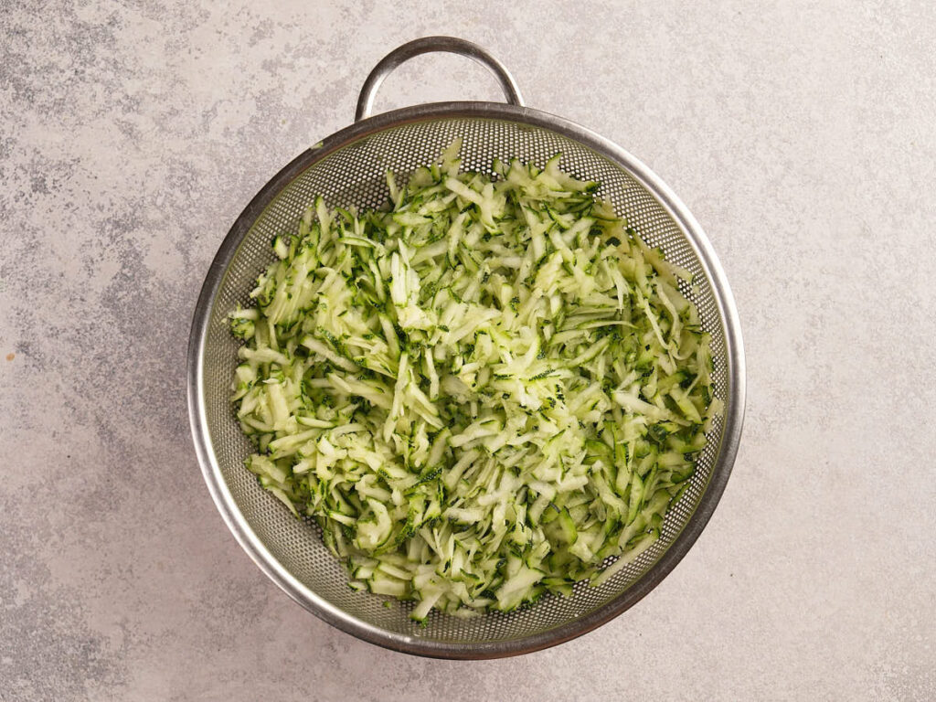 A metal colander filled with shredded cucumber sits on a light gray textured surface.