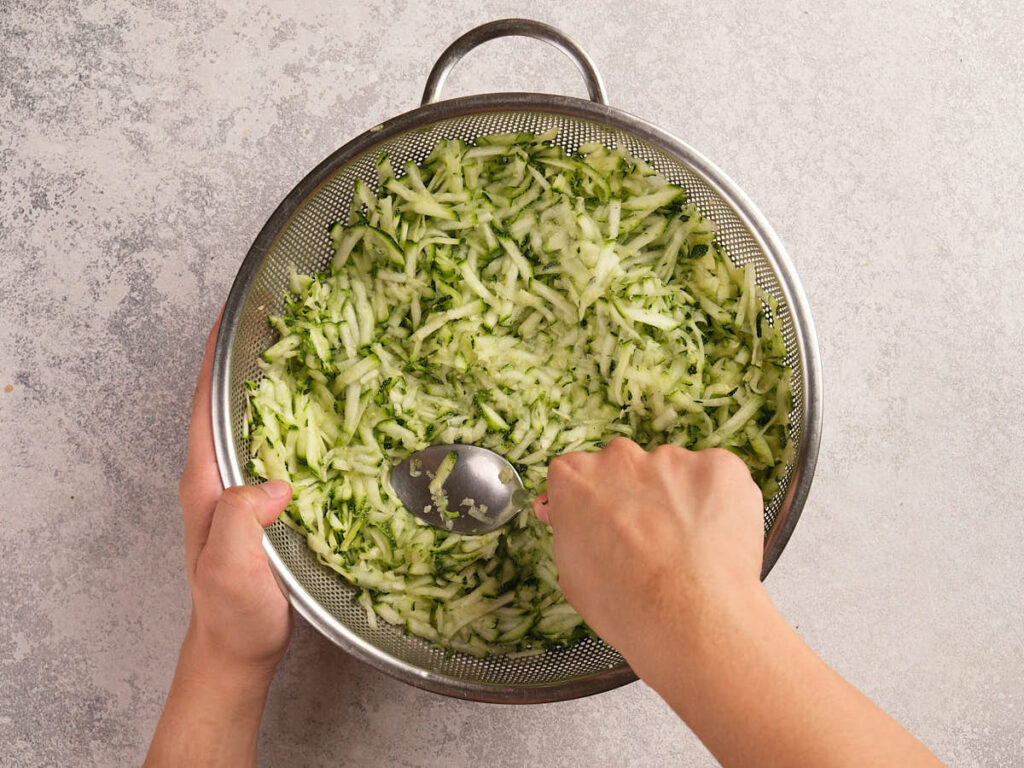 A person uses a spoon to press shredded cucumber in a metal colander over a light-colored countertop.