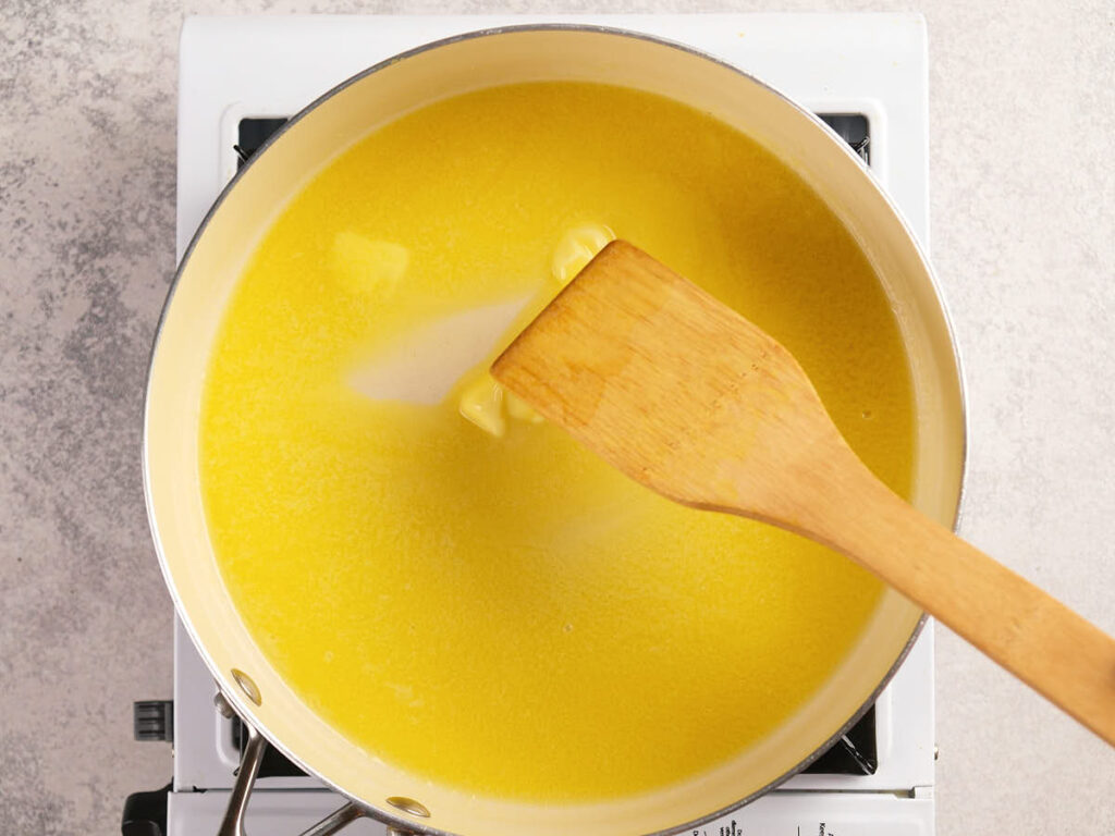 A wooden spatula stirs melting butter in a saucepan on a stovetop.