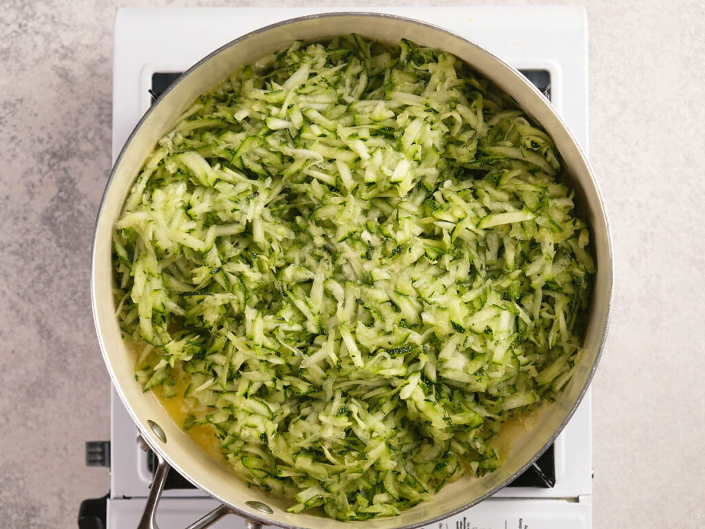 A metal pan filled with shredded zucchini sits on a stovetop, viewed from above.