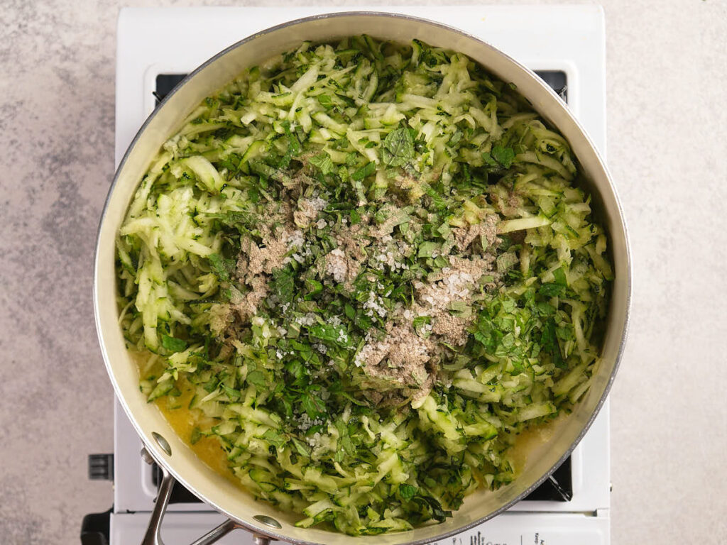 A metal pan on a stove contains shredded zucchini mixed with chopped herbs, seasonings, and salt, ready to be cooked.